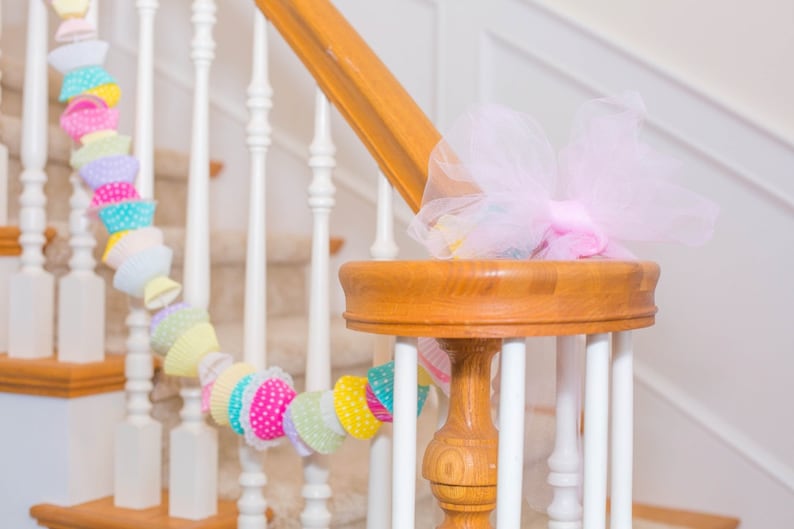 May include: A white staircase with a wooden handrail decorated with a colorful garland of paper cupcake liners and a pink tulle bow.