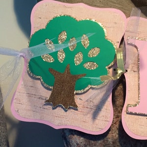 May include: A pink and green banner with a green tree with a brown trunk and gold glitter. The tree is on a pink background with a white and gold glitter border.