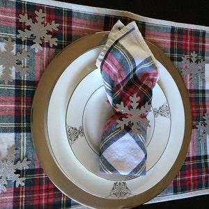 Op de afbeelding: Een wit en rood geruit placemat met een zilveren onderbord en een wit dinerbord. Een witte servet met een rood en zwart geruit ontwerp is gevouwen en op het onderbord gelegd. Er zijn zilveren sneeuwvlokdecoraties verspreid over de placemat.