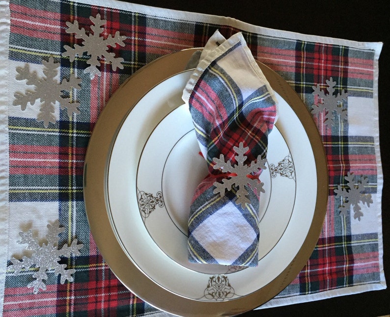 Op de afbeelding: Een wit en rood geruit placemat met een zilveren onderbord en een wit dinerbord. Een witte servet met een rood en zwart geruit ontwerp is gevouwen en op het onderbord gelegd. Er zijn zilveren sneeuwvlokdecoraties verspreid over de placemat.