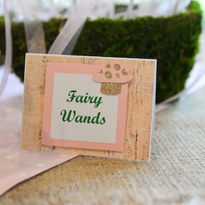 May include: A pink and brown sign with the text "Fairy Wands" in green lettering. A small, glittery, pink and white mushroom is on the sign.