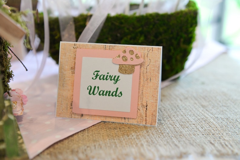 May include: A pink and brown sign with the text "Fairy Wands" in green lettering. A small, glittery, pink and white mushroom is on the sign.