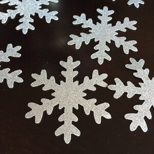 May include: A collection of silver glitter snowflakes on a dark brown surface.