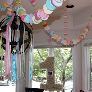May include: A black metal chandelier with colorful ribbons hanging from it. The chandelier is decorated with pastel-colored paper cupcake liners, creating a festive atmosphere.