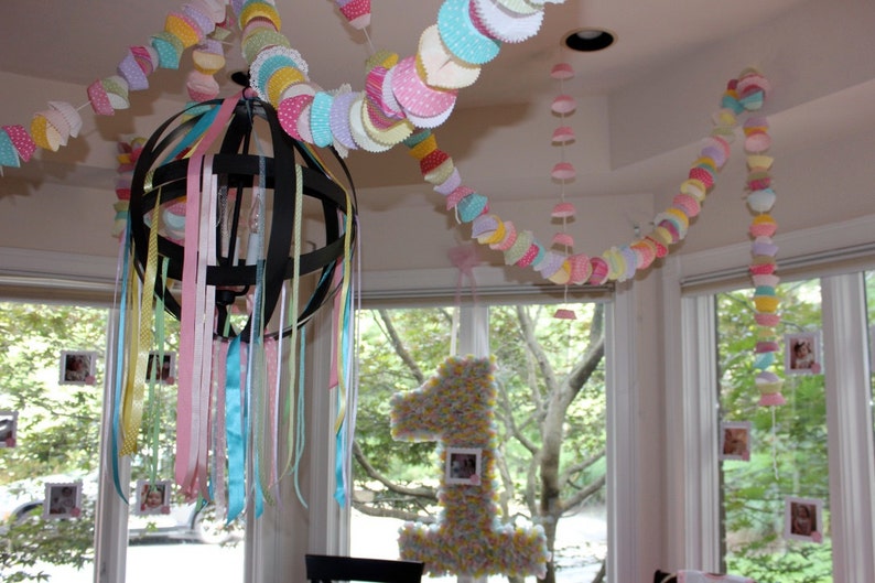 May include: A black metal chandelier with colorful ribbons hanging from it. The chandelier is decorated with pastel-colored paper cupcake liners, creating a festive atmosphere.