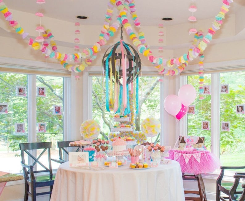 May include: A pink and white birthday party table setting with a cake, cupcakes, and other treats. The table is decorated with pink and white balloons, and a banner that says "One".