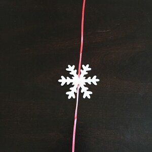 Op de afbeelding: Een witte sneeuwvlok ornament met een rood lint eraan bevestigd. De sneeuwvlok hangt tegen een donkerbruine achtergrond.