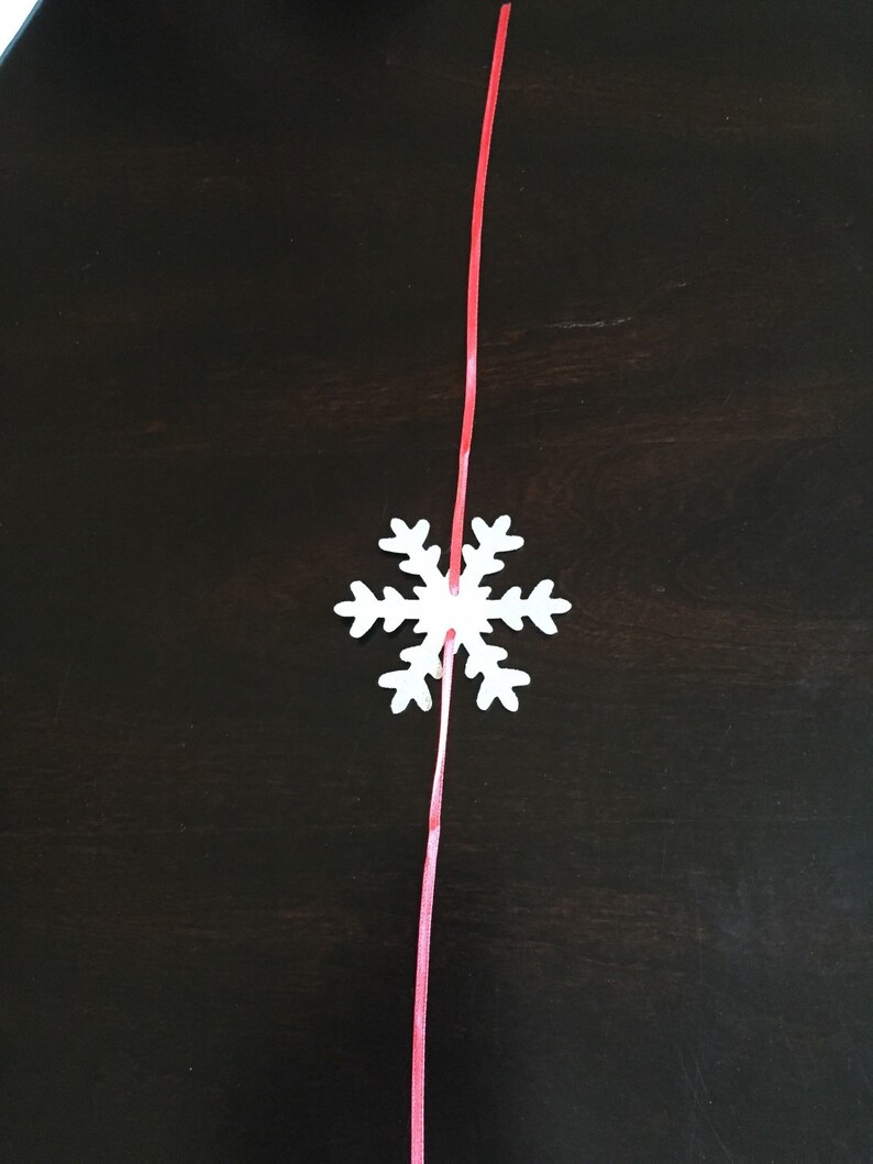 Op de afbeelding: Een witte sneeuwvlok ornament met een rood lint eraan bevestigd. De sneeuwvlok hangt tegen een donkerbruine achtergrond.