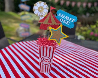 May include: A festive circus-themed centerpiece featuring a popcorn container with "POP CORN" text, surrounded by glittery cutouts of a Ferris wheel, circus tent, star, and an "ADMIT ONE" ticket. The centerpiece sits on a red and white striped tablecloth.