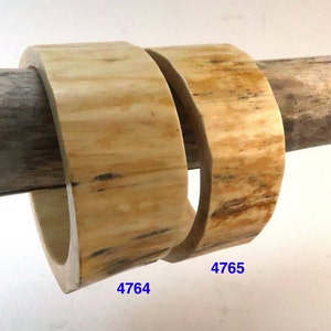 May include: Two square-shaped, light-colored bangles with natural wood grain patterns. The bangles are displayed on a wooden dowel. The numbers "4764" and "4765" are printed in blue on the bangles.