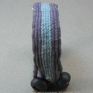 May include: A headband made of braided rope with a blue and purple striped design. The headband has two dark blue pom-poms on the ends.