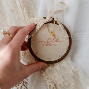 May include: A circular wooden embroidery hoop containing cream-coloured fabric with the embroidered name Matilda in pink thread. A small gold star charm hangs from a thin cord at the top, and a cream lace ribbon is tied around the hoop frame.