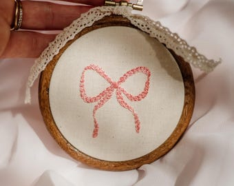 May include: A circular embroidery hoop with a pink bow design stitched on cream fabric. The wooden frame has a metal clasp and is trimmed with lace. The background is a soft, white fabric. The hoop is a decorative item.