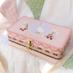 May include: A pink rectangular box with a gold clasp and a decorative border. The box features a painted illustration of a goose, daisies, and strawberries, with the name "Daisy" written on it. A gold charm hangs from the clasp.