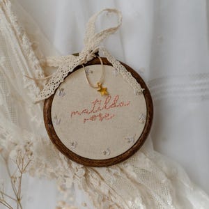 May include: A circular wooden embroidery hoop containing cream-coloured fabric with the embroidered name matilda rose in pink thread. Small floral accents surround the text. A gold star charm hangs from a thin cord, and a cream lace ribbon is tied.