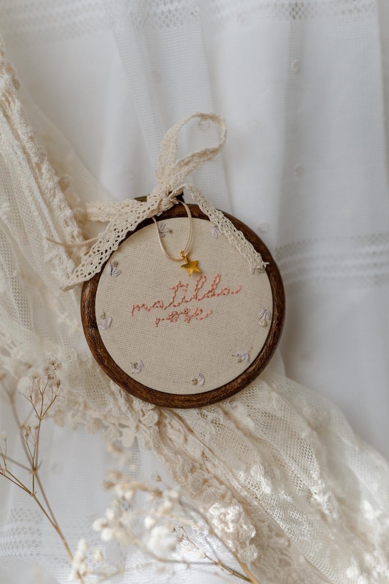 May include: A circular wooden embroidery hoop containing cream-coloured fabric with the embroidered name matilda rose in pink thread. Small floral accents surround the text. A gold star charm hangs from a thin cord, and a cream lace ribbon is tied.