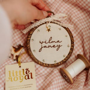 May include: A circular wooden embroidery hoop frame containing off-white fabric with the embroidered name "wilma janey" and small floral motifs. The hoop features a cream lace bow, a gold star charm, and a tag labelled "little royal".