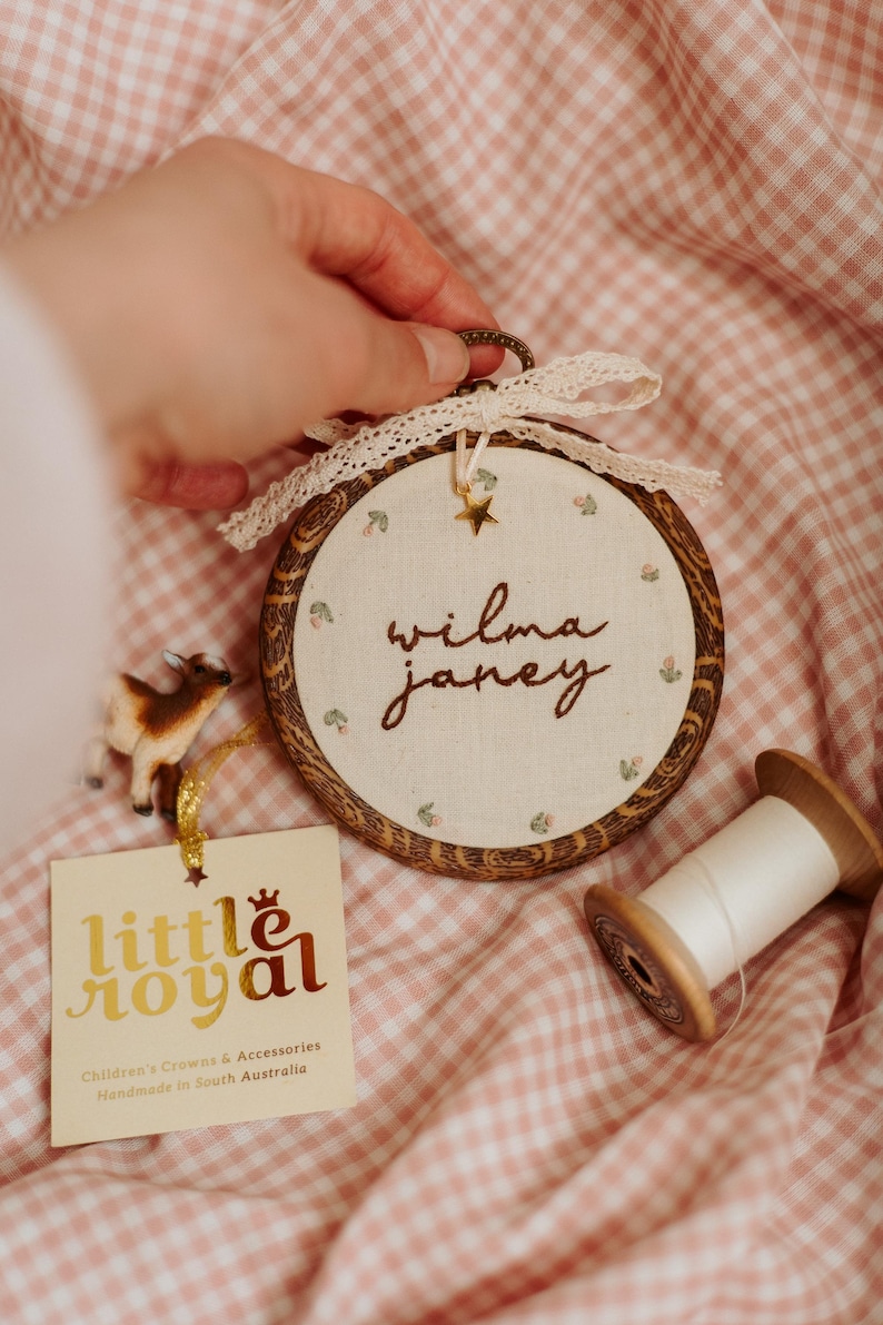 May include: A circular wooden embroidery hoop frame containing off-white fabric with the embroidered name "wilma janey" and small floral motifs. The hoop features a cream lace bow, a gold star charm, and a tag labelled "little royal".
