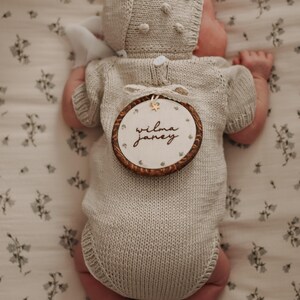 May include: A beige knitted short-sleeve romper and matching bonnet with bobble details. A round wooden embroidery hoop is placed on the back, featuring the name "wilma janey" embroidered in black script with a small gold star accent.