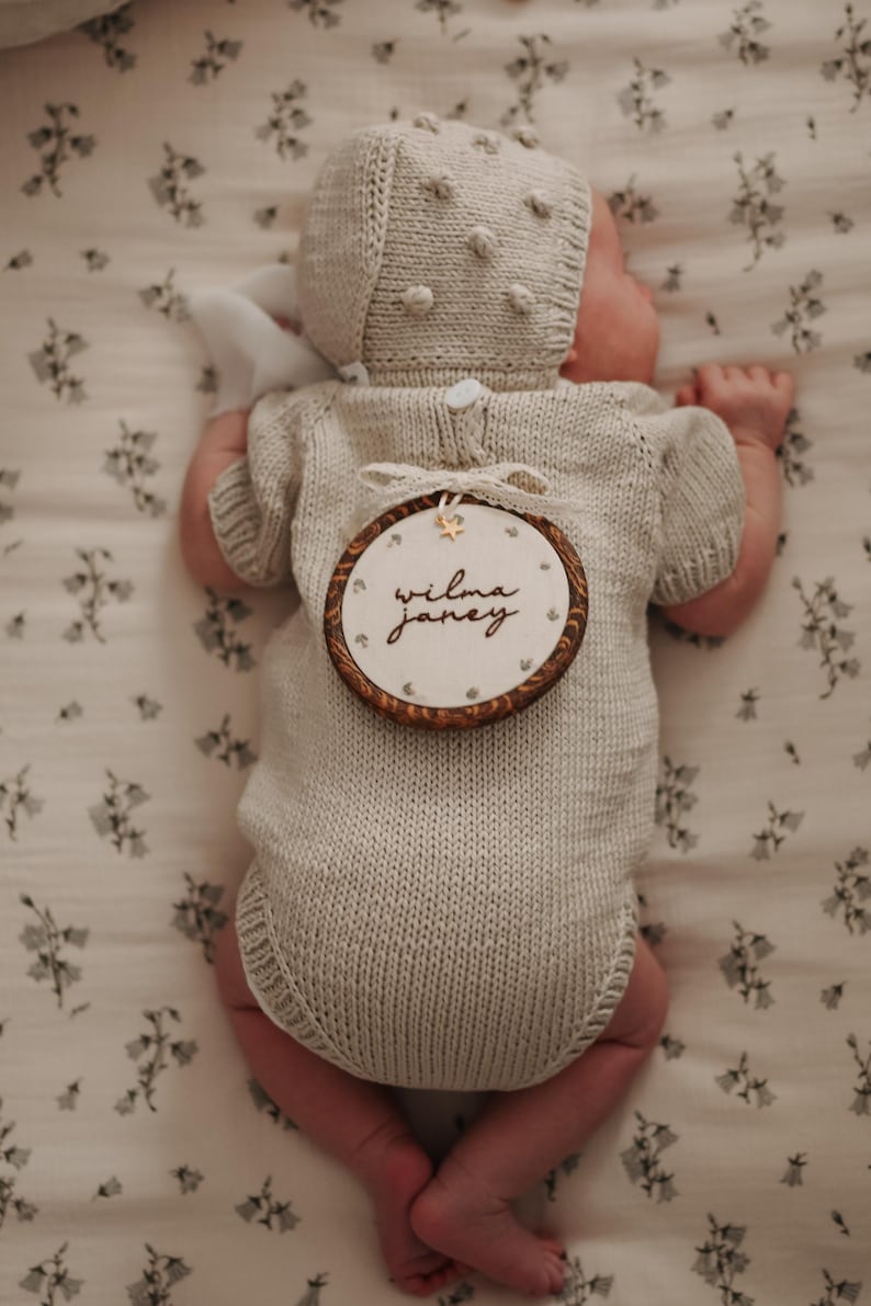 May include: A beige knitted short-sleeve romper and matching bonnet with bobble details. A round wooden embroidery hoop is placed on the back, featuring the name "wilma janey" embroidered in black script with a small gold star accent.