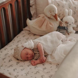 May include: A sleeping infant wearing a beige knit bonnet with small pom-poms and a matching knit top, wrapped in a white muslin swaddle. The infant lies on a cream-coloured sheet with a small floral pattern, surrounded by a plush swan, a bear, and a rabbit.