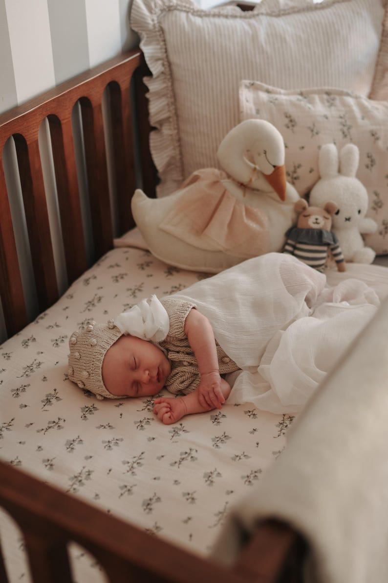 May include: A sleeping infant wearing a beige knit bonnet with small pom-poms and a matching knit top, wrapped in a white muslin swaddle. The infant lies on a cream-coloured sheet with a small floral pattern, surrounded by a plush swan, a bear, and a rabbit.