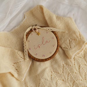 May include: A circular wooden embroidery hoop containing beige fabric with the name Isla stitched in pink thread, surrounded by small pink floral accents. A gold star charm hangs from a cream lace ribbon tied to the top of the hoop.