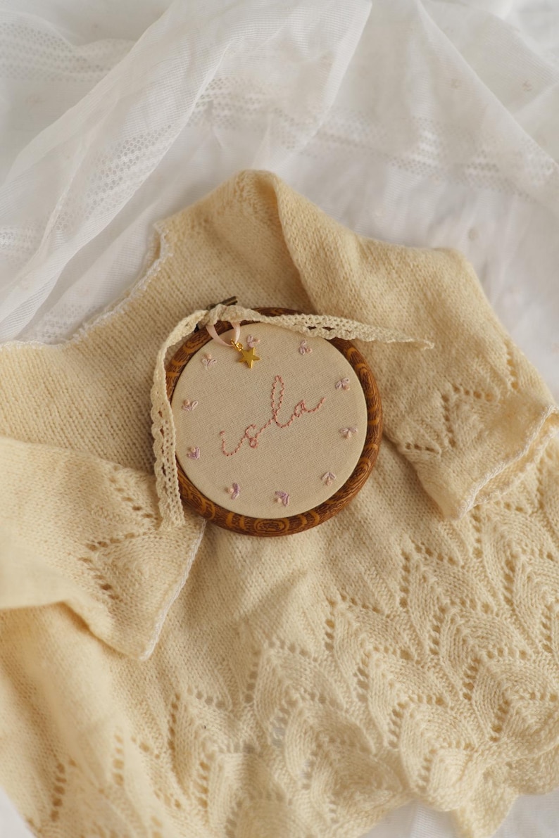 May include: A circular wooden embroidery hoop containing beige fabric with the name Isla stitched in pink thread, surrounded by small pink floral accents. A gold star charm hangs from a cream lace ribbon tied to the top of the hoop.