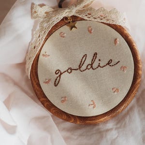 May include: A circular wooden embroidery hoop containing cream-coloured fabric with the word goldie stitched in brown thread. Small pink floral accents surround the text. A cream lace ribbon and a small gold star charm are attached to the top.