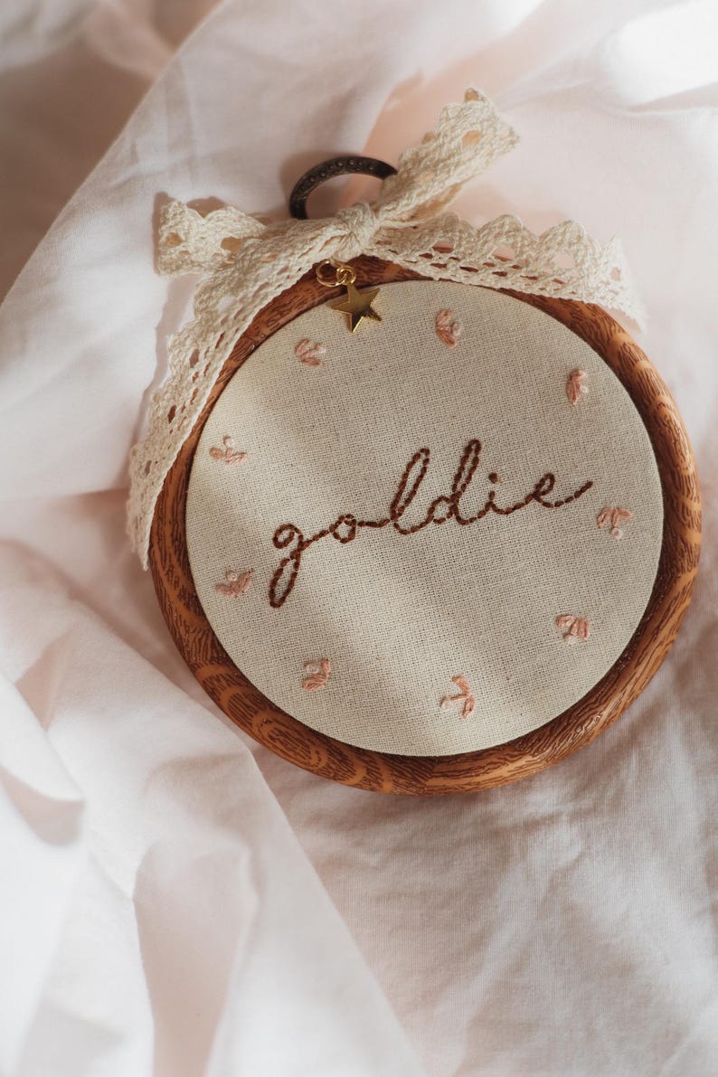 May include: A circular wooden embroidery hoop containing cream-coloured fabric with the word goldie stitched in brown thread. Small pink floral accents surround the text. A cream lace ribbon and a small gold star charm are attached to the top.