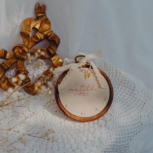 May include: A circular wooden embroidery hoop containing cream-coloured fabric with embroidered text that reads "matilda rose". A small gold star charm hangs from a cream lace ribbon tied at the top of the hoop, which is positioned near a gold bow.