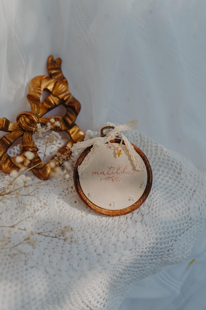 May include: A circular wooden embroidery hoop containing cream-coloured fabric with embroidered text that reads "matilda rose". A small gold star charm hangs from a cream lace ribbon tied at the top of the hoop, which is positioned near a gold bow.