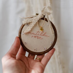 May include: A circular wooden embroidery hoop containing cream-coloured fabric with the embroidered name "matilda rose" in pink thread. Small floral motifs surround the text, and a gold star charm hangs from a cream lace ribbon tied at the top.