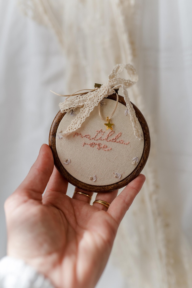May include: A circular wooden embroidery hoop containing cream-coloured fabric with the embroidered name "matilda rose" in pink thread. Small floral motifs surround the text, and a gold star charm hangs from a cream lace ribbon tied at the top.