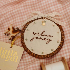 May include: A circular wooden embroidery hoop containing cream fabric with the embroidered name "wilma janey" in brown thread, accented by small floral motifs, a gold star charm, and a cream lace bow. A small goat figurine and tag are present.