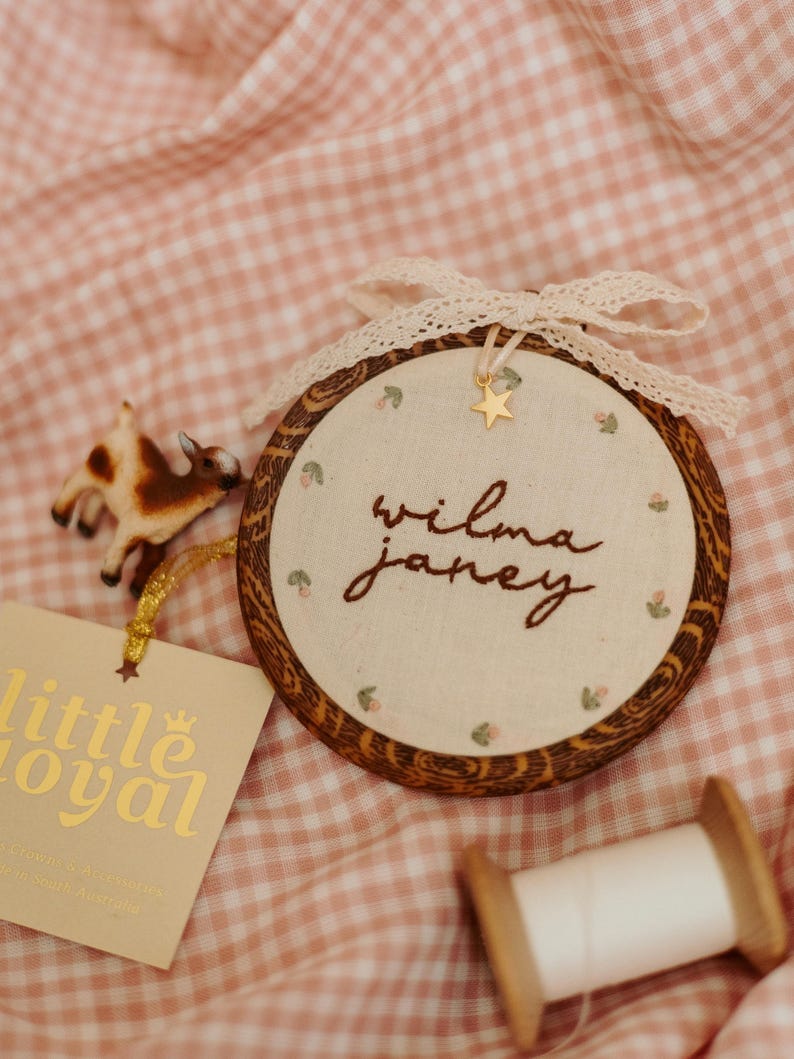 May include: A circular wooden embroidery hoop containing cream fabric with the embroidered name "wilma janey" in brown thread, accented by small floral motifs, a gold star charm, and a cream lace bow. A small goat figurine and tag are present.