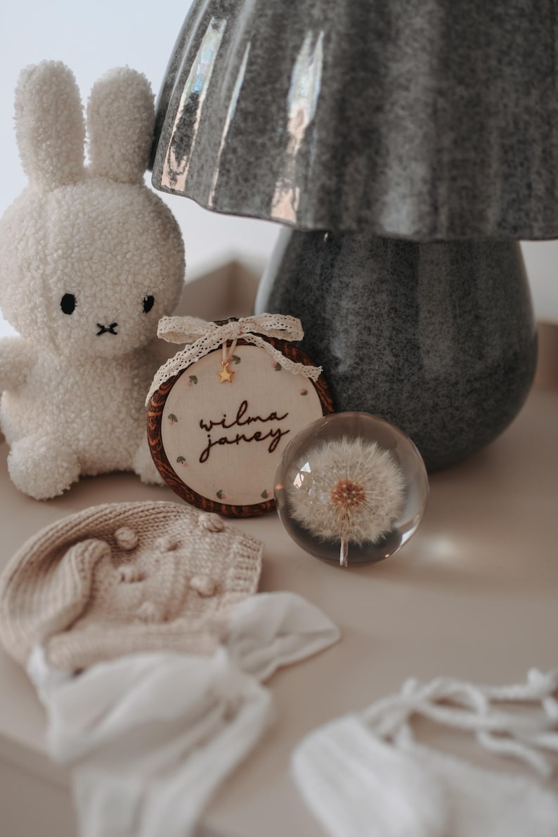 May include: A white plush rabbit toy, a circular wooden embroidery hoop with the name Wilma Janey stitched in black thread, a clear glass sphere containing a dandelion seed head, and a beige knitted bonnet with white ribbon ties.