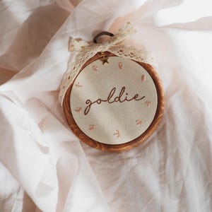 May include: A circular wooden embroidery hoop containing off-white fabric with the word goldie stitched in brown thread. Small pink floral accents surround the text. A cream-coloured lace ribbon is tied at the top with a small gold star charm.