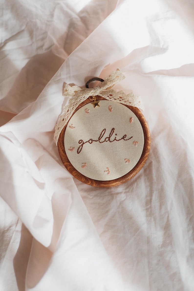 May include: A circular wooden embroidery hoop containing off-white fabric with the word goldie stitched in brown thread. Small pink floral accents surround the text. A cream-coloured lace ribbon is tied at the top with a small gold star charm.