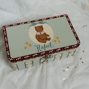 Op de afbeelding: Een rechthoekige, lichtblauwe decoratieve doos met een scharnierend deksel. De doos heeft een geschilderde illustratie van een teddybeer, sterren en de naam "Rafael". De randen zijn afgewerkt met een donkerrood en wit patroon en de doos is beveiligd met een goudkleurige sluiting.