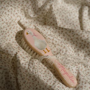 Puede incluir: Un cepillo de bebé de madera con un mango rosa y una ilustración pintada de un ganso con un lazo rosa. El cepillo tiene cerdas blancas y la palabra "Magnolia" está escrita en el mango en cursiva. Se adjunta una cinta de encaje blanco.