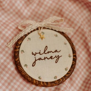 May include: A circular embroidery hoop with a dark wood grain frame containing off-white fabric. The text "wilma janey" is embroidered in brown thread, surrounded by small floral motifs. A cream lace ribbon and a small gold star charm are attached.