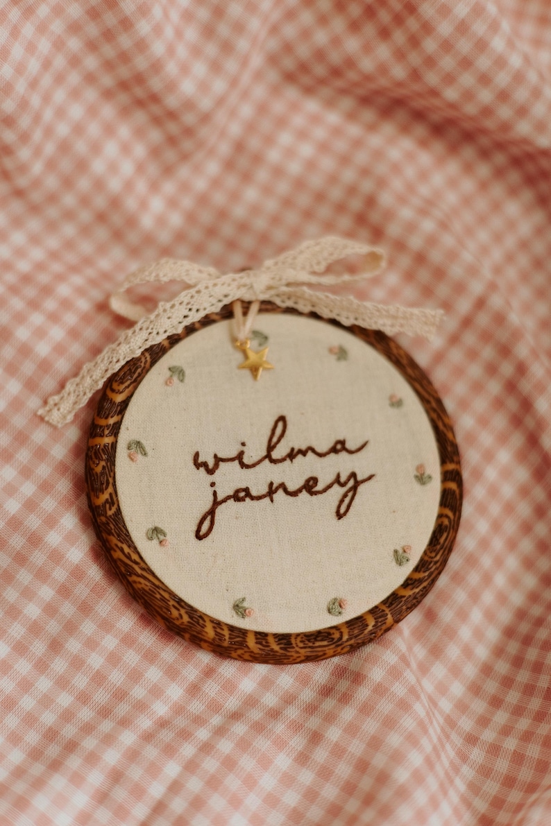 May include: A circular embroidery hoop with a dark wood grain frame containing off-white fabric. The text "wilma janey" is embroidered in brown thread, surrounded by small floral motifs. A cream lace ribbon and a small gold star charm are attached.