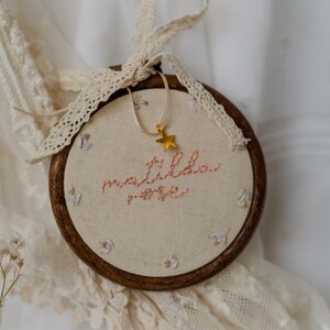 May include: A circular wooden embroidery hoop containing off-white fabric with the name Matilda Rose stitched in pink thread. Small floral motifs are embroidered around the text. A gold star charm hangs from a cream ribbon tied to the top.