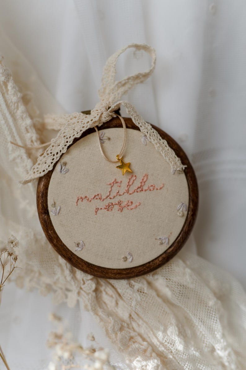 May include: A circular wooden embroidery hoop containing off-white fabric with the name Matilda Rose stitched in pink thread. Small floral motifs are embroidered around the text. A gold star charm hangs from a cream ribbon tied to the top.