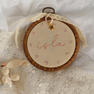 May include: A circular wooden embroidery hoop containing beige fabric with the name Isla stitched in pink thread. The design features small embroidered pink flowers, a gold star charm hanging from a ribbon, and a cream-coloured lace trim border.