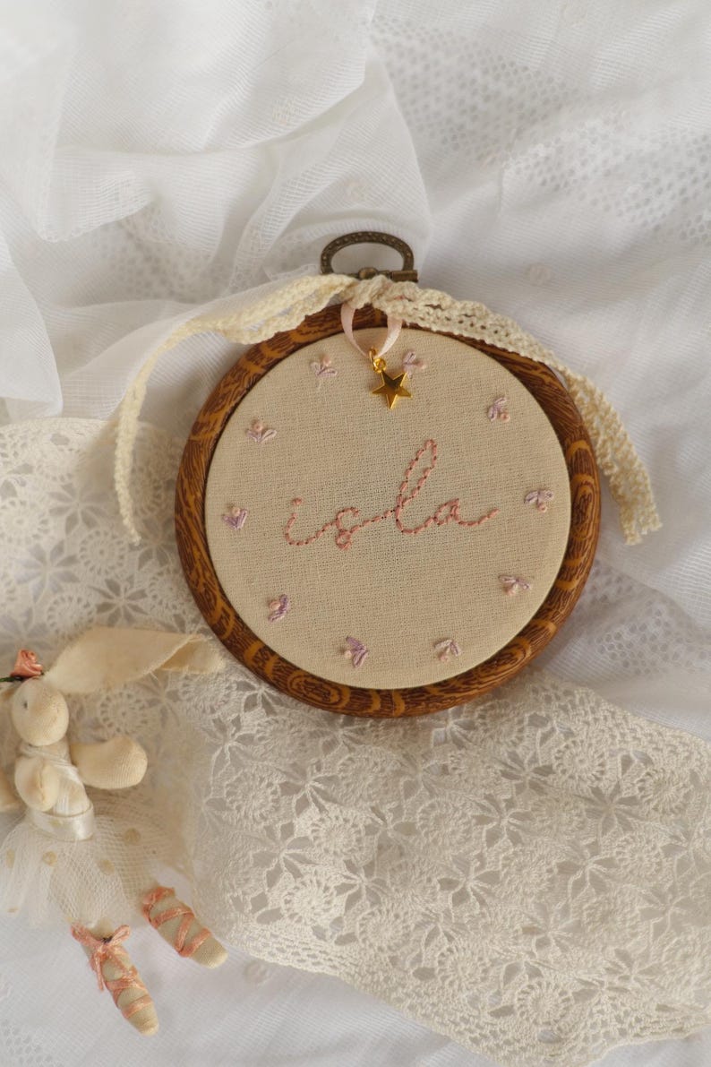 May include: A circular wooden embroidery hoop containing beige fabric with the name Isla stitched in pink thread. The design features small embroidered pink flowers, a gold star charm hanging from a ribbon, and a cream-coloured lace trim border.