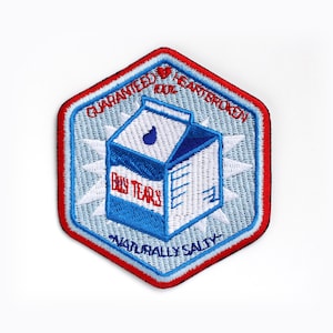 Op de afbeelding: Een blauw en wit geborduurde patch in de vorm van een zeshoek met de tekst "Guaranteed to make you heartbroken" en "Baby Tears Naturally Salty" rond een cartoon afbeelding van een melkpak.