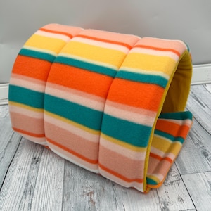 May include: A cylindrical pet tunnel made of soft fleece fabric featuring horizontal stripes in vibrant shades of orange, teal, yellow, and light pink. The interior of the tunnel is lined with a solid mustard yellow fabric, creating a cozy space.