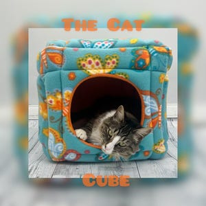 May include: A teal fleece cat cube featuring a colorful butterfly pattern and an orange circular entrance. The text THE CAT CUBE is printed in orange above and below the cube. A brown and white tabby cat rests inside the soft fabric structure.
